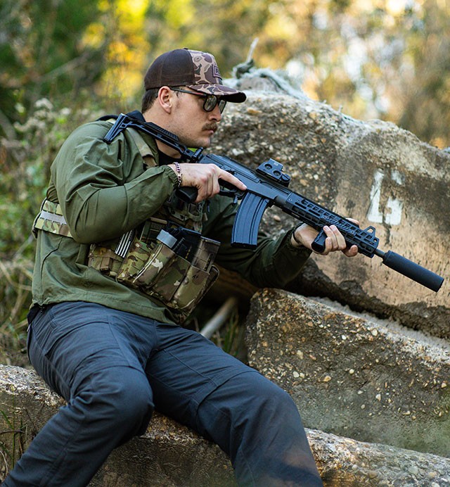 Shooter in multicam kneeling on rocks with a suppressed rifle